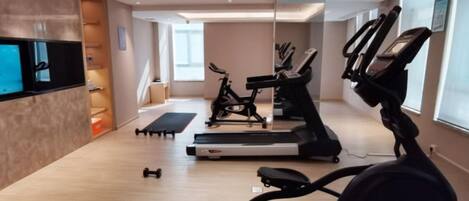 A modern gym with cardio equipment, a flat-screen TV, and a large window providing natural light.