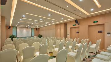Un salón de conferencias con filas de sillas blancas, una pantalla de proyector y un escenario.