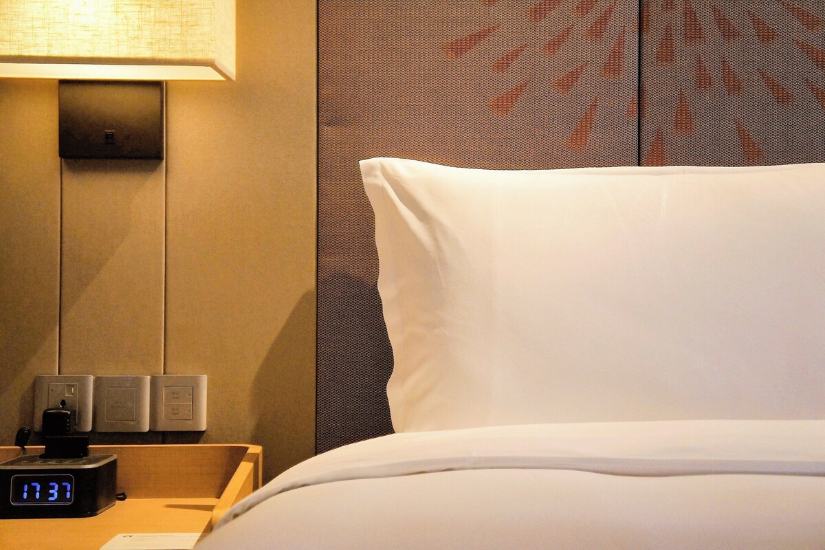 A hotel room with a bed, a digital clock, a lamp, and a wall with a patterned design.