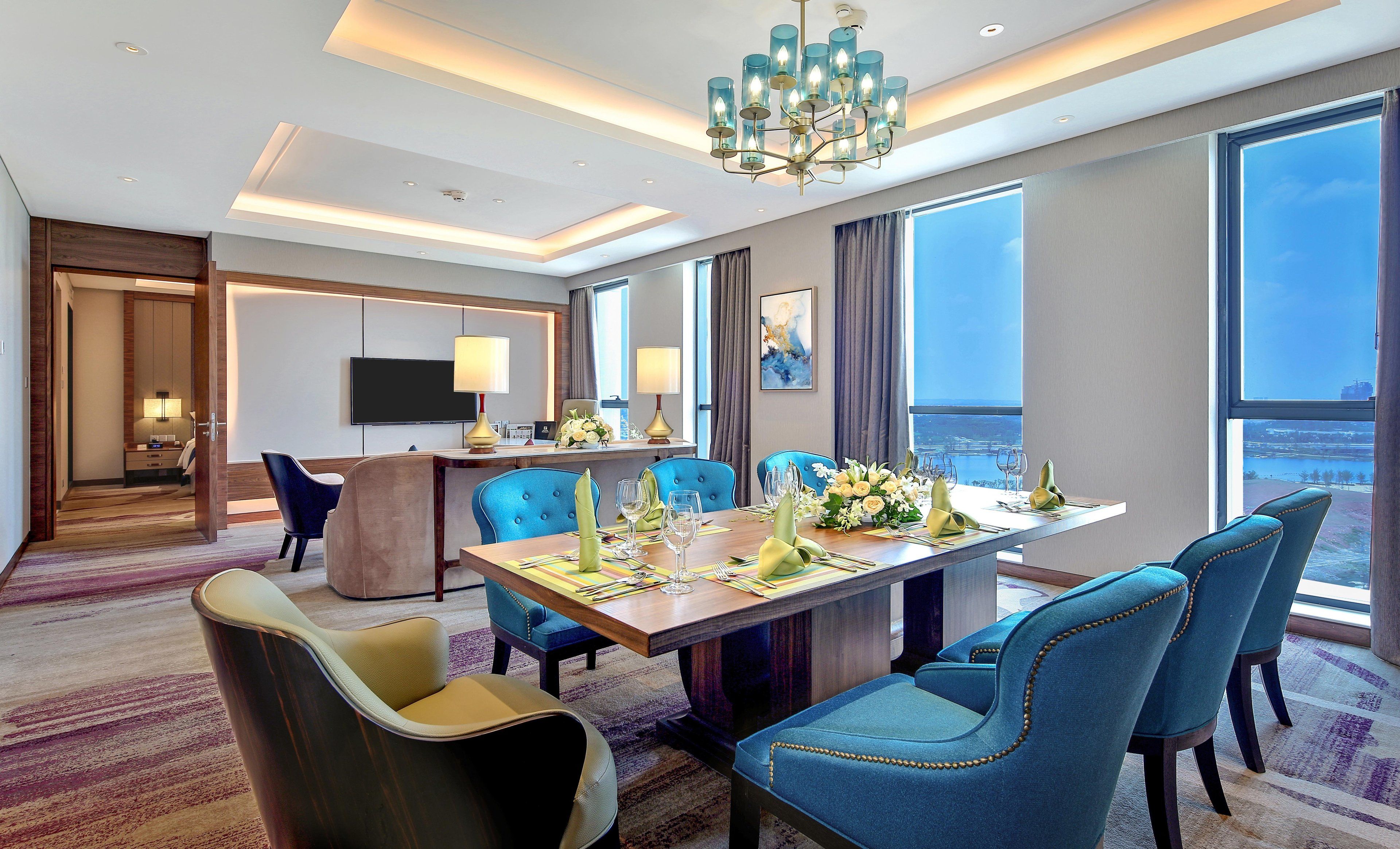 Una moderna sala da pranzo di un hotel con un tavolo grande imbandito per sei persone, sedie rivestite di blu, un lampadario e vista sulla città attraverso ampie finestre.