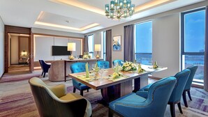 A modern hotel dining room with a large table set for six, blue upholstered chairs, a chandelier, and a view of the city through large windows.