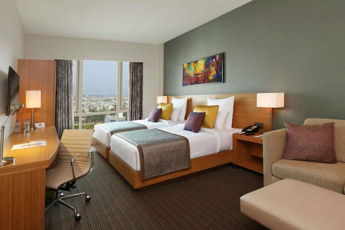 Habitación de hotel con cama, dos mesitas de noche, escritorio, silla, sofá y un ventanal con vistas a la ciudad.