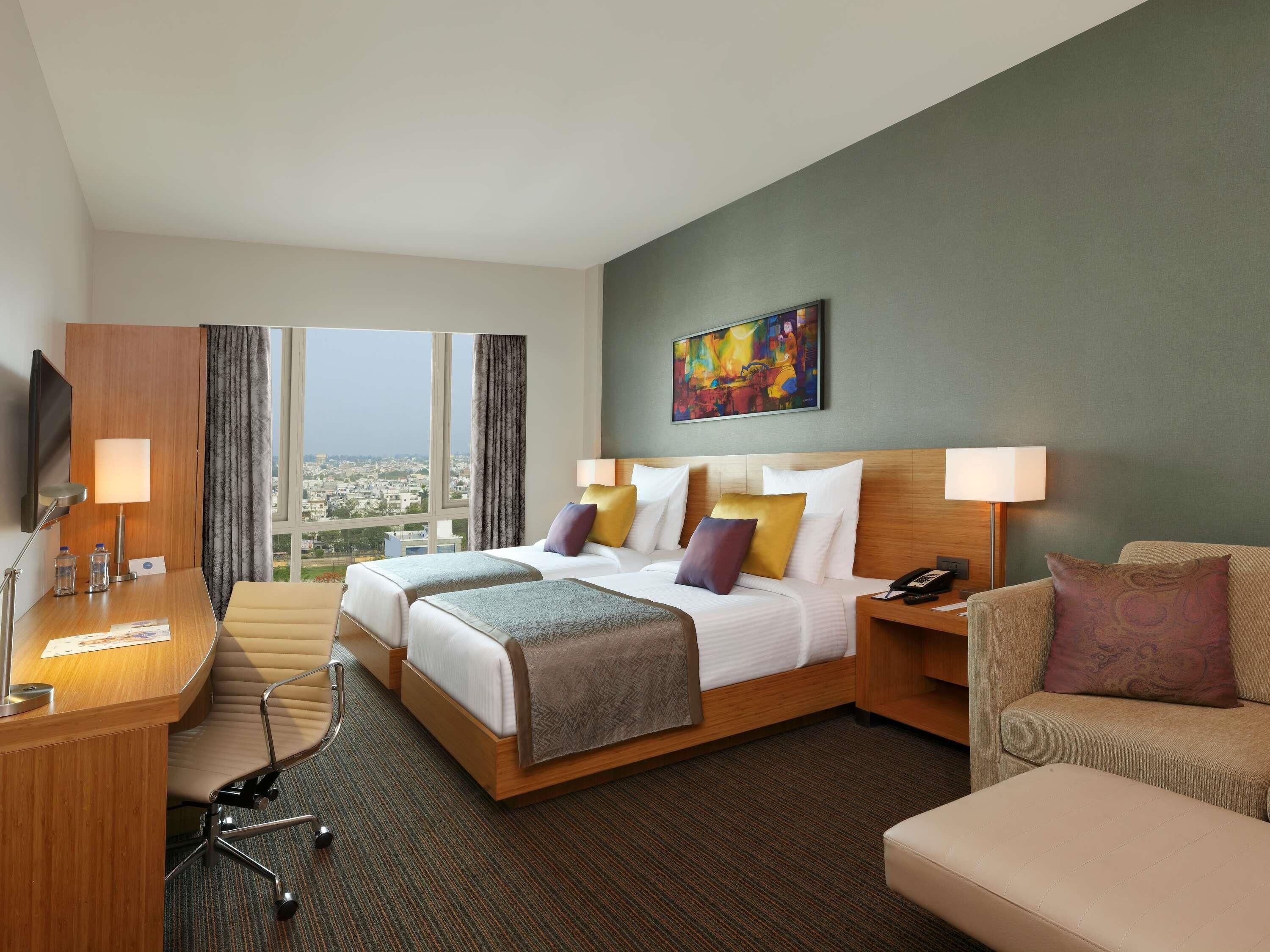 Habitación de hotel con cama, dos mesitas de noche, escritorio, silla, sofá y un ventanal con vistas a la ciudad.
