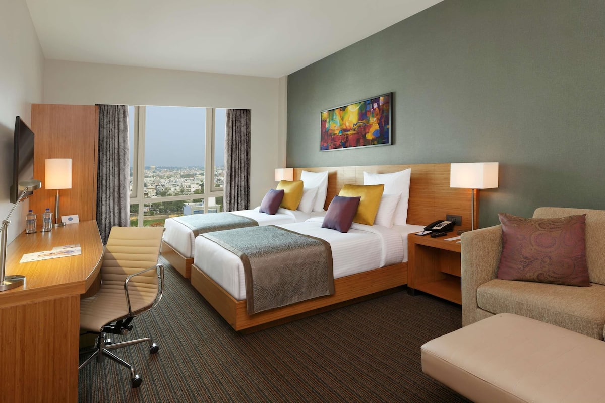 Habitación de hotel con cama, dos mesitas de noche, escritorio, silla, sofá y un ventanal con vistas a la ciudad.