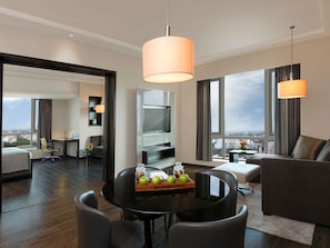 A modern hotel room with a dining area, a sofa, a TV, and a view of the city.