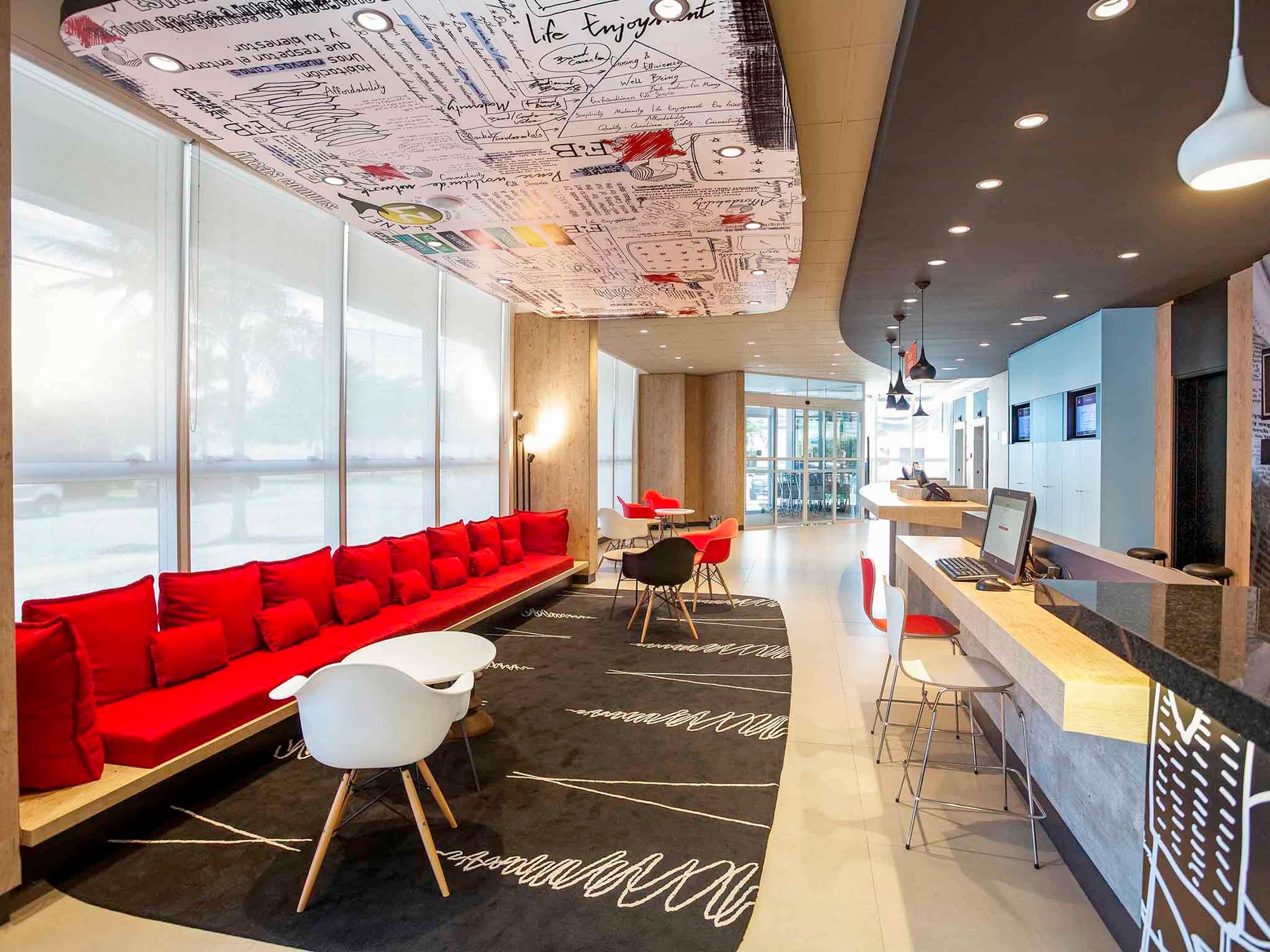 Un vestíbulo de hotel moderno con un sofá rojo, sillas blancas, un mostrador de recepción y ventanas amplias.