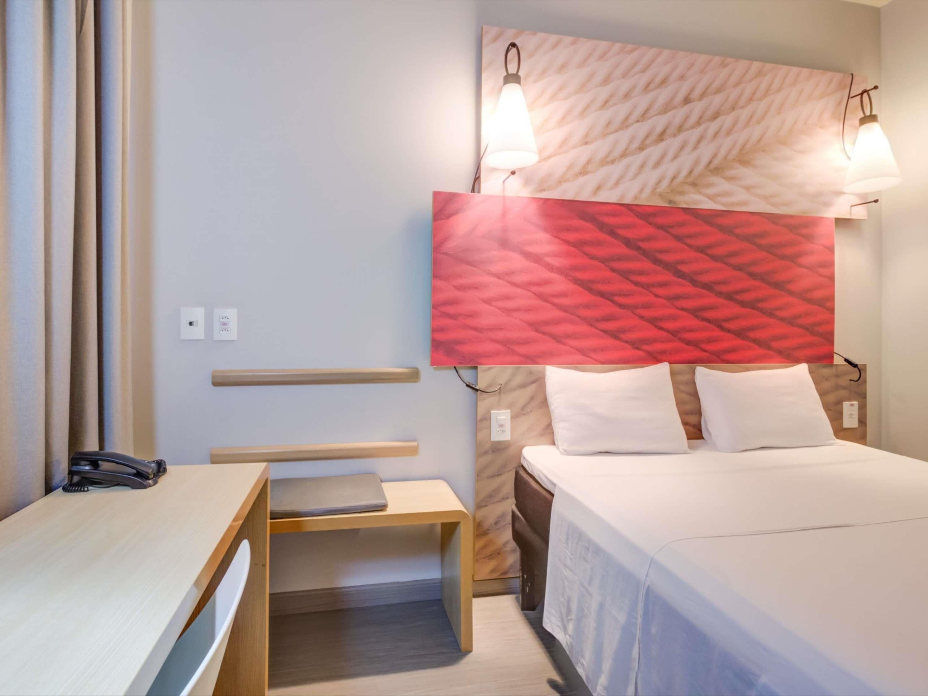 A hotel room with a bed, bedside tables, a desk, and a headboard with wall-mounted lights.