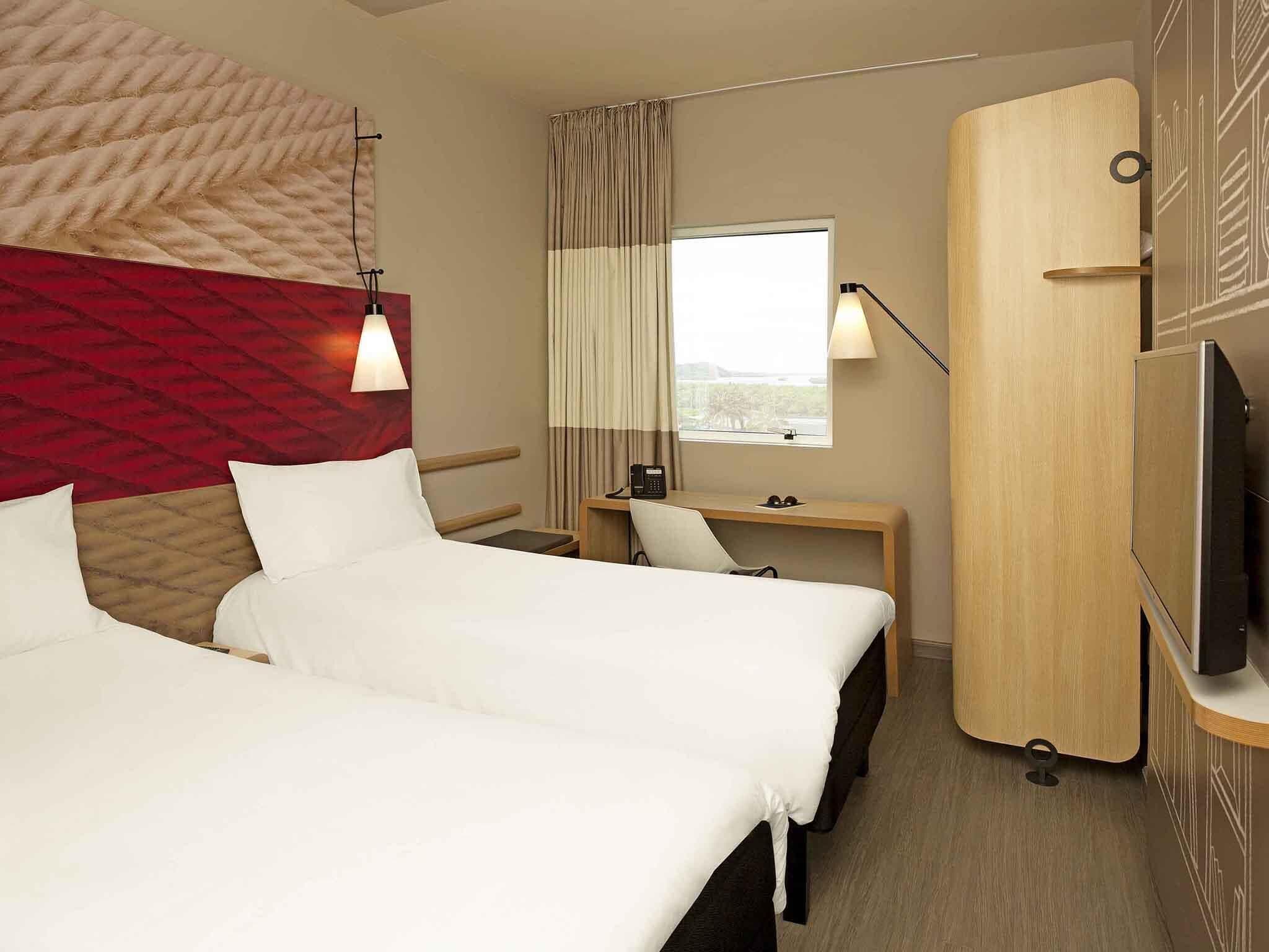 A hotel room with two beds, a desk, a chair, a TV, a window with curtains, and a wall-mounted lamp.