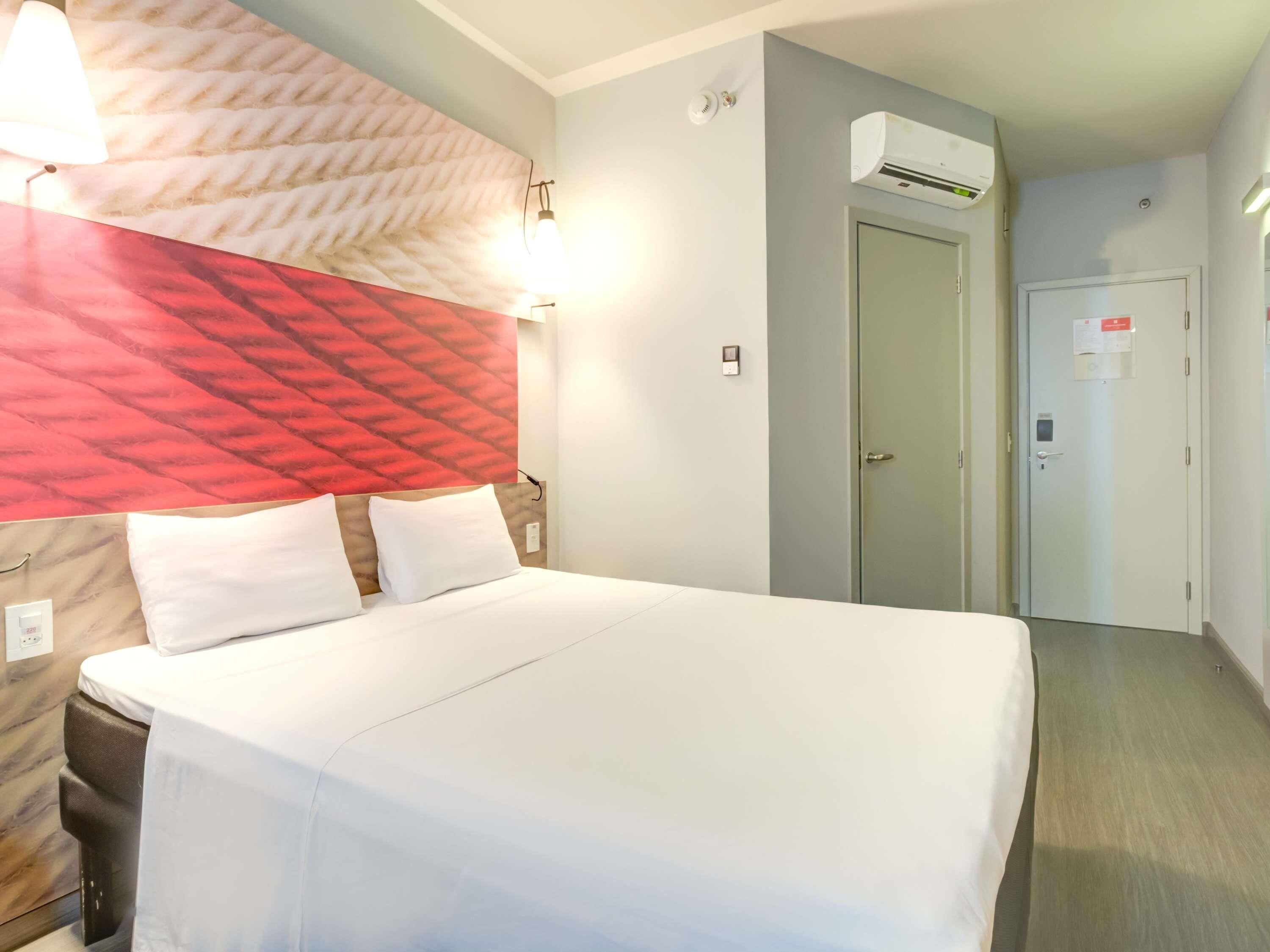 A hotel room with a bed, a headboard with a red and beige pattern, an air conditioning unit, a door with a sign, and a wall-mounted light fixture.
