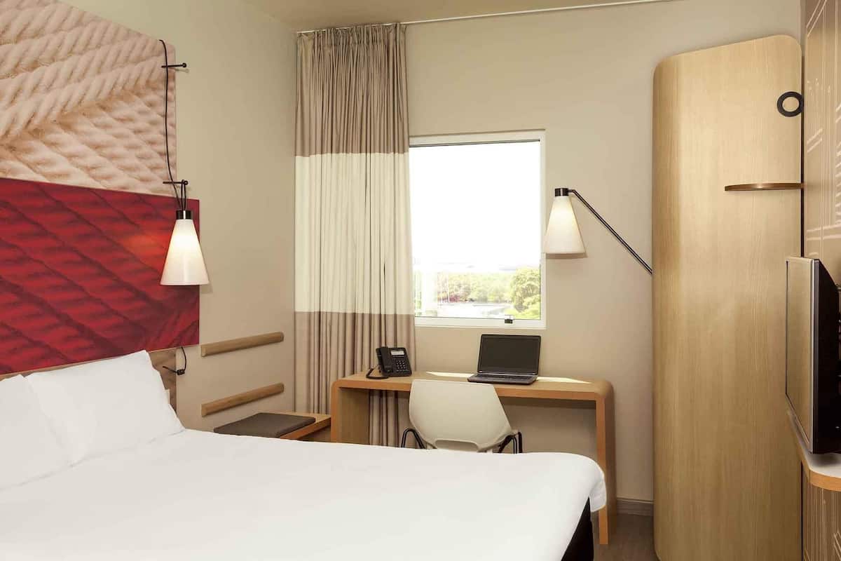 A hotel room with a bed, a desk with a laptop, a telephone, a chair, a wardrobe, and a window with curtains.