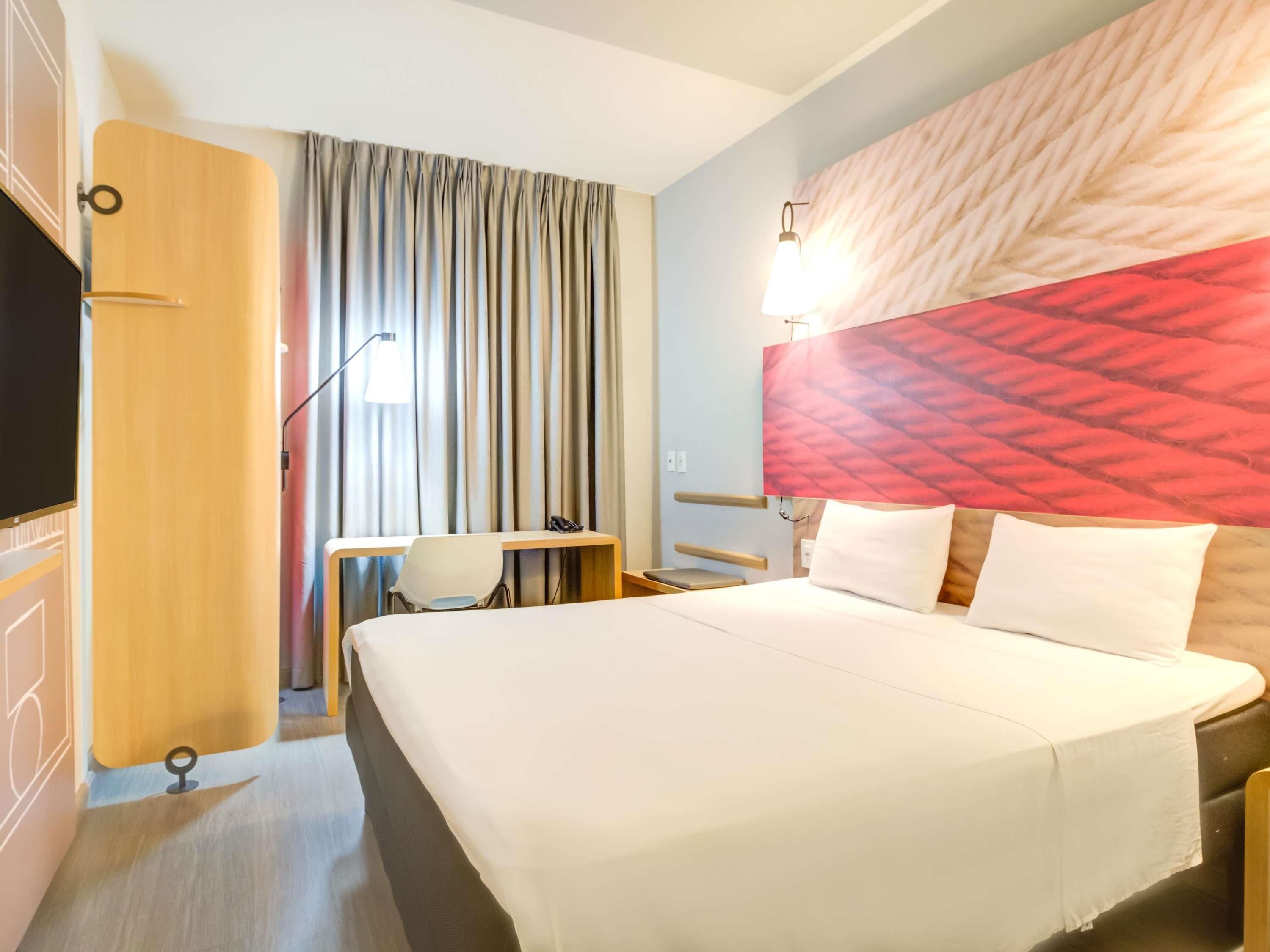 Quarto de hotel com cama, escrivaninha, cadeira, televisão e cortina.