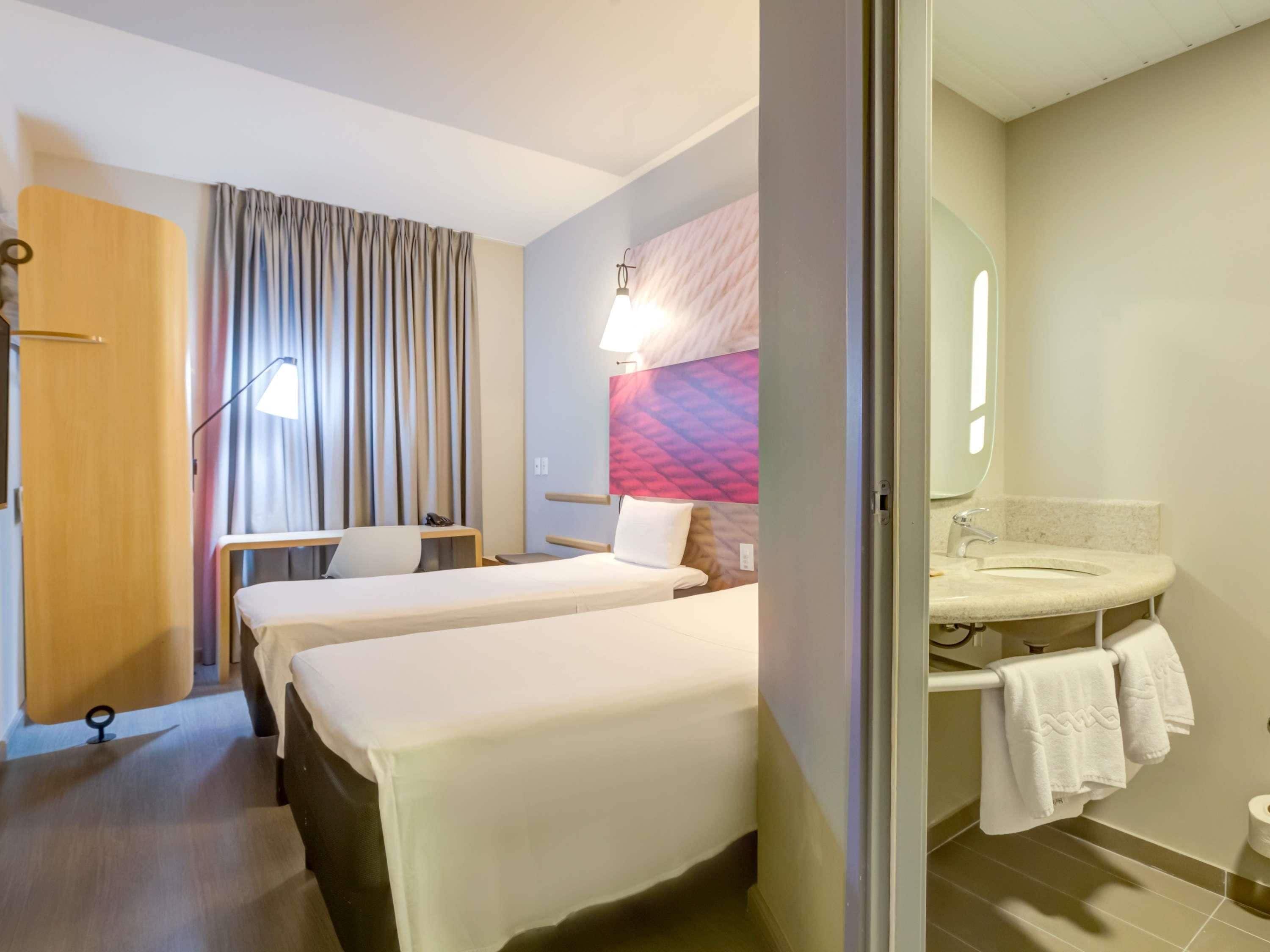 A hotel room with two beds, a desk, a chair, a desk lamp, a wall-mounted TV, and a bathroom with a sink and towel rack.