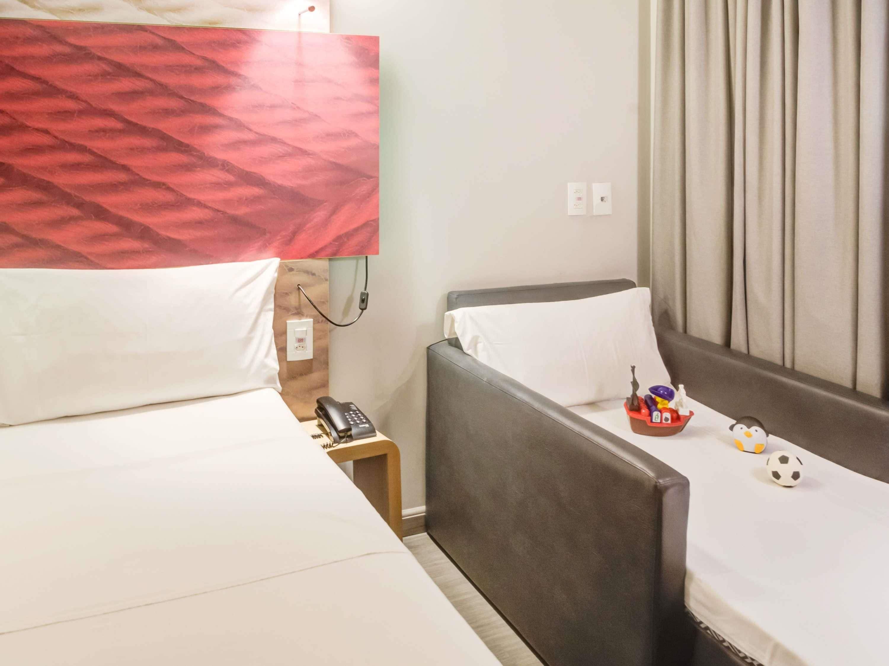 Quarto de hotel com cama, sofá, mesa de cabeceira com telefone e pequena mesa com um boneco de pinguim e uma bola de futebol.
