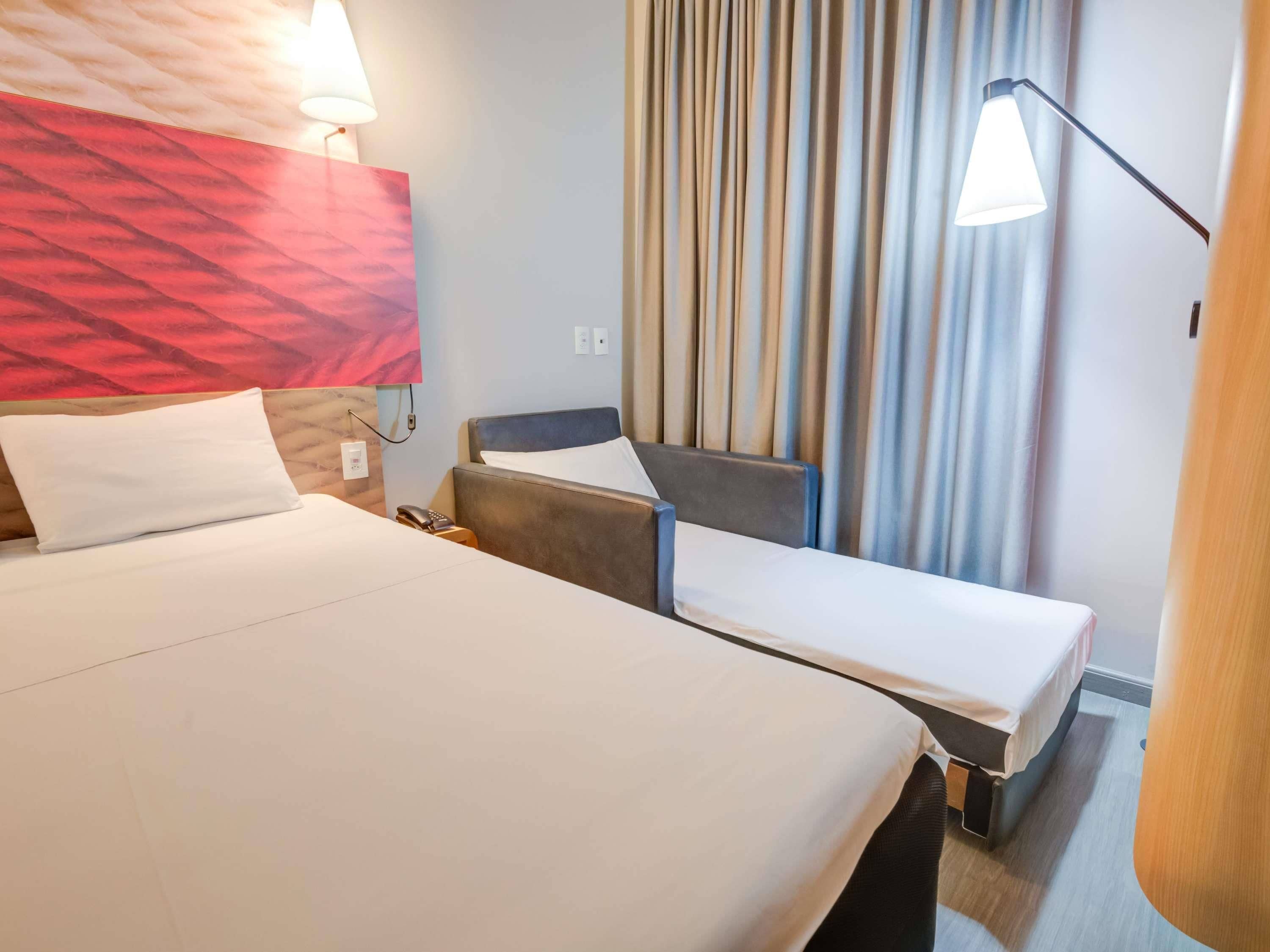A hotel room with a large bed, a desk, and a chair. There is a lamp on the desk and a phone on the nightstand.