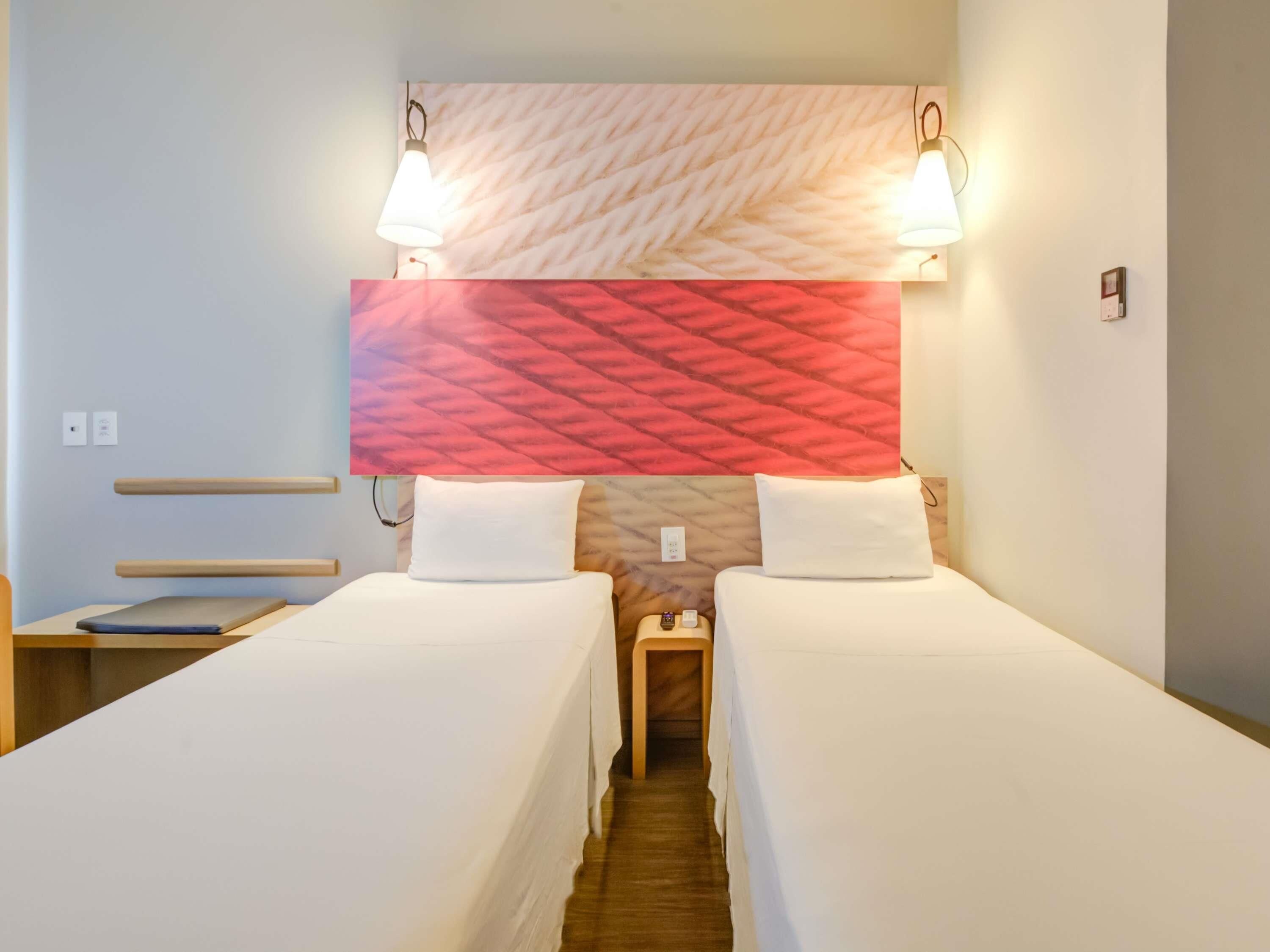 A hotel room with two single beds, a wooden bench, a nightstand with a lamp, and a patterned headboard.