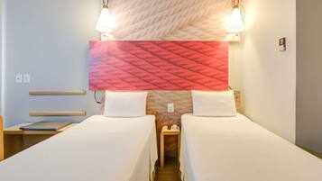 A hotel room with two single beds, a wooden bench, a nightstand with a lamp, and a patterned headboard.