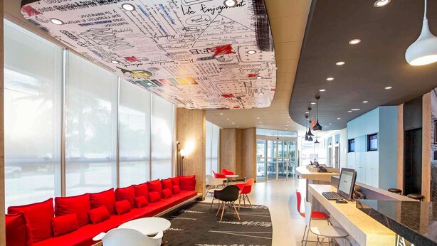 Un vestíbulo de hotel moderno con un sofá rojo, sillas blancas, un mostrador de recepción y ventanas amplias.