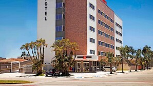 Un moderno edificio de hotel con un llamativo logo 'ibis', varios pisos y un estacionamiento con coches y palmeras.