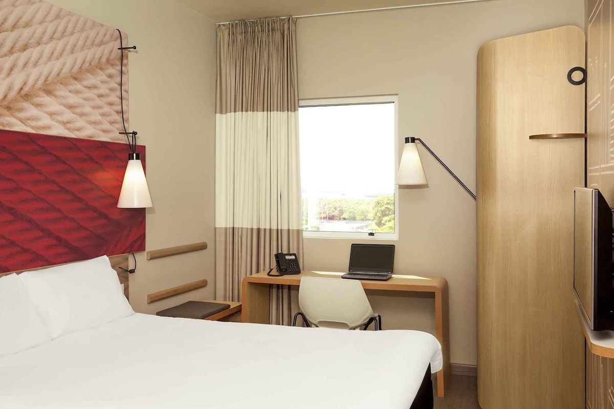 A hotel room with a bed, a desk with a laptop, a telephone, a chair, a wardrobe, and a window with curtains.