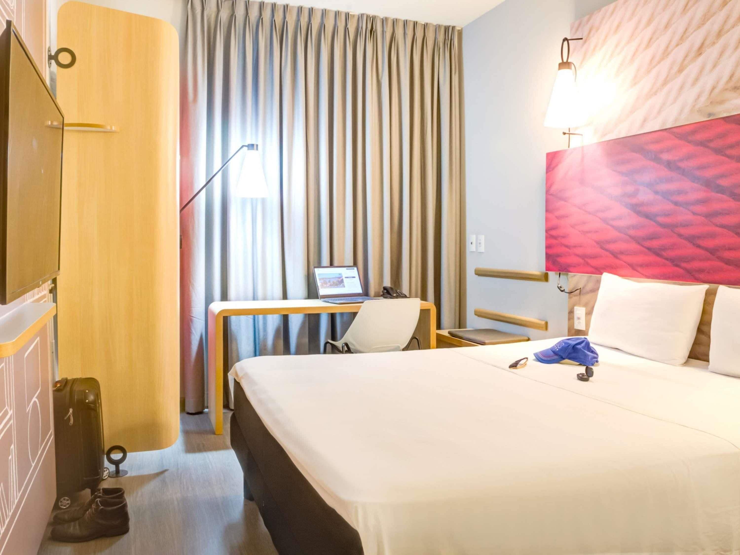 A hotel room with a bed, a desk with a laptop, a chair, a suitcase, and a pair of boots.