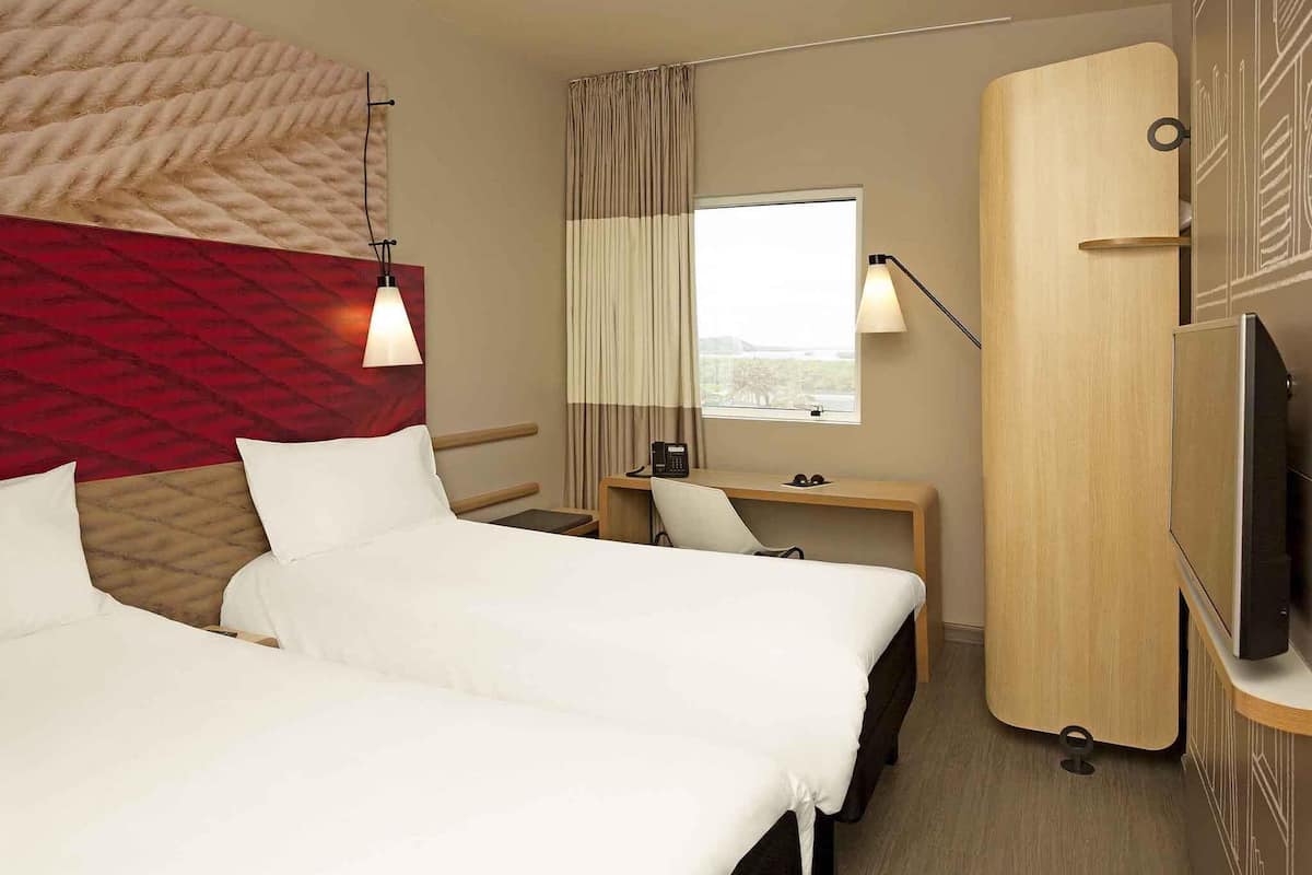 A hotel room with two beds, a desk, a chair, a TV, a window with curtains, and a wall-mounted lamp.