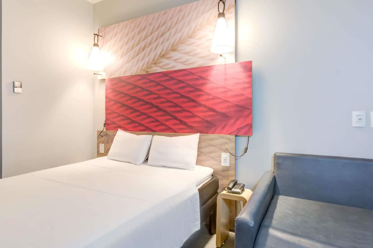 A hotel room with a bed, a desk, a chair, a wall-mounted light, and a power outlet.