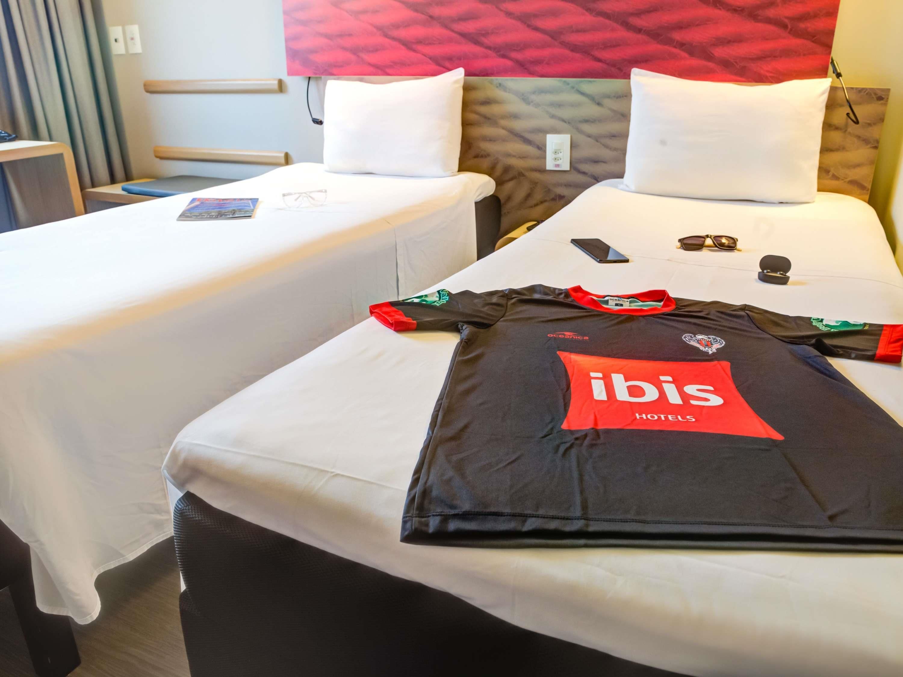 Quarto de hotel com duas camas de solteiro, uma camisola vermelha e preta na cama, um par de óculos de sol, um telemóvel e uma pequena caixa.