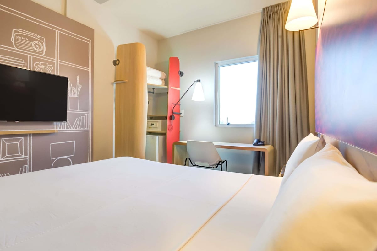 Habitación de hotel con cama, escritorio, silla, televisor y ventana con cortinas.