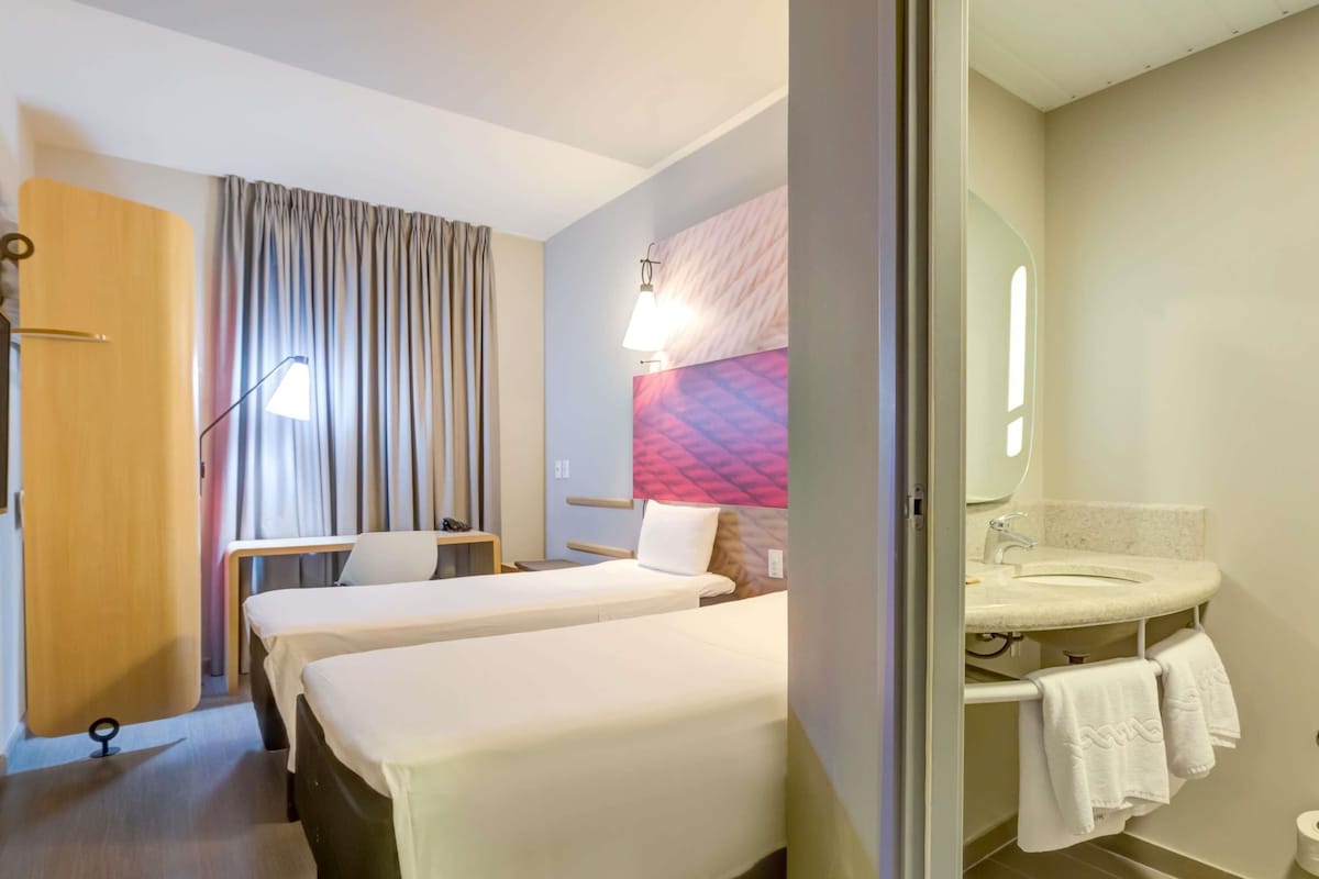 Habitación de hotel con dos camas, un escritorio, una silla, una lámpara de escritorio, televisor montado en la pared y baño con lavamanos y toallero.