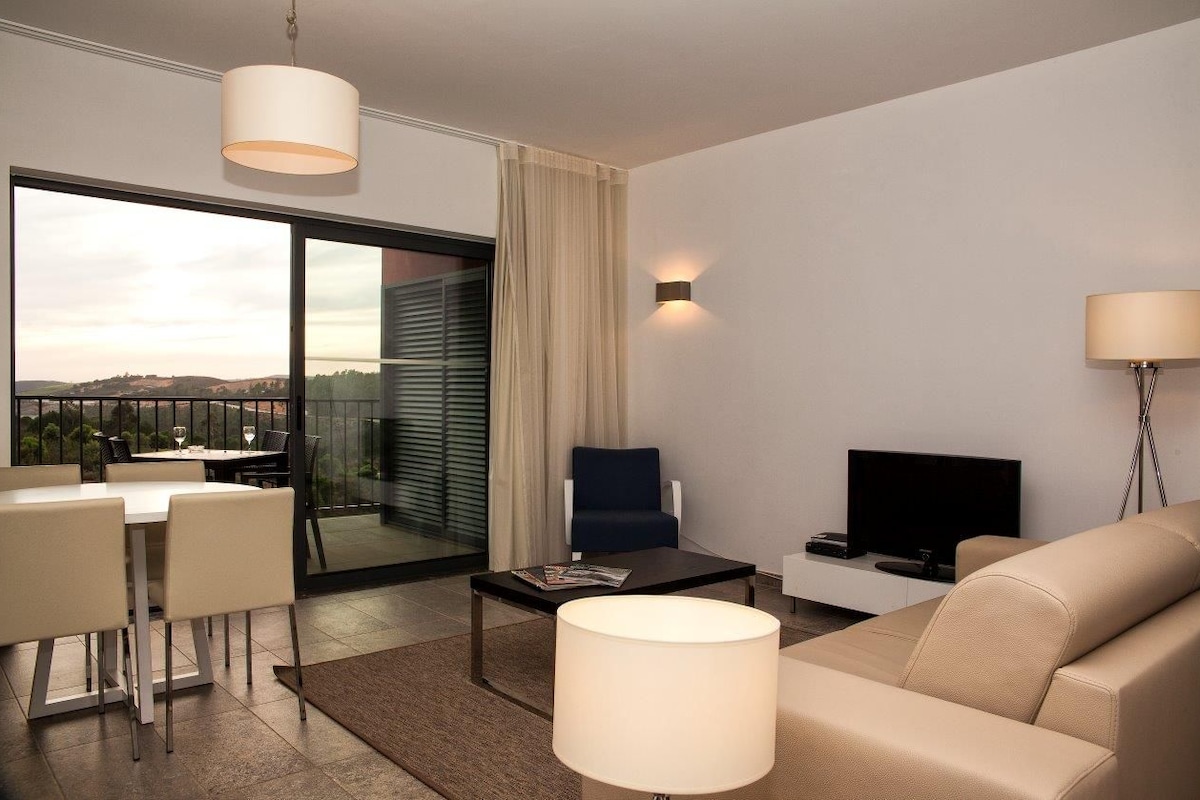 Une chambre d’hôtel moderne dotée d’un coin repas, d’un canapé, d’une télévision et d’une grande fenêtre offrant une vue dégagée.