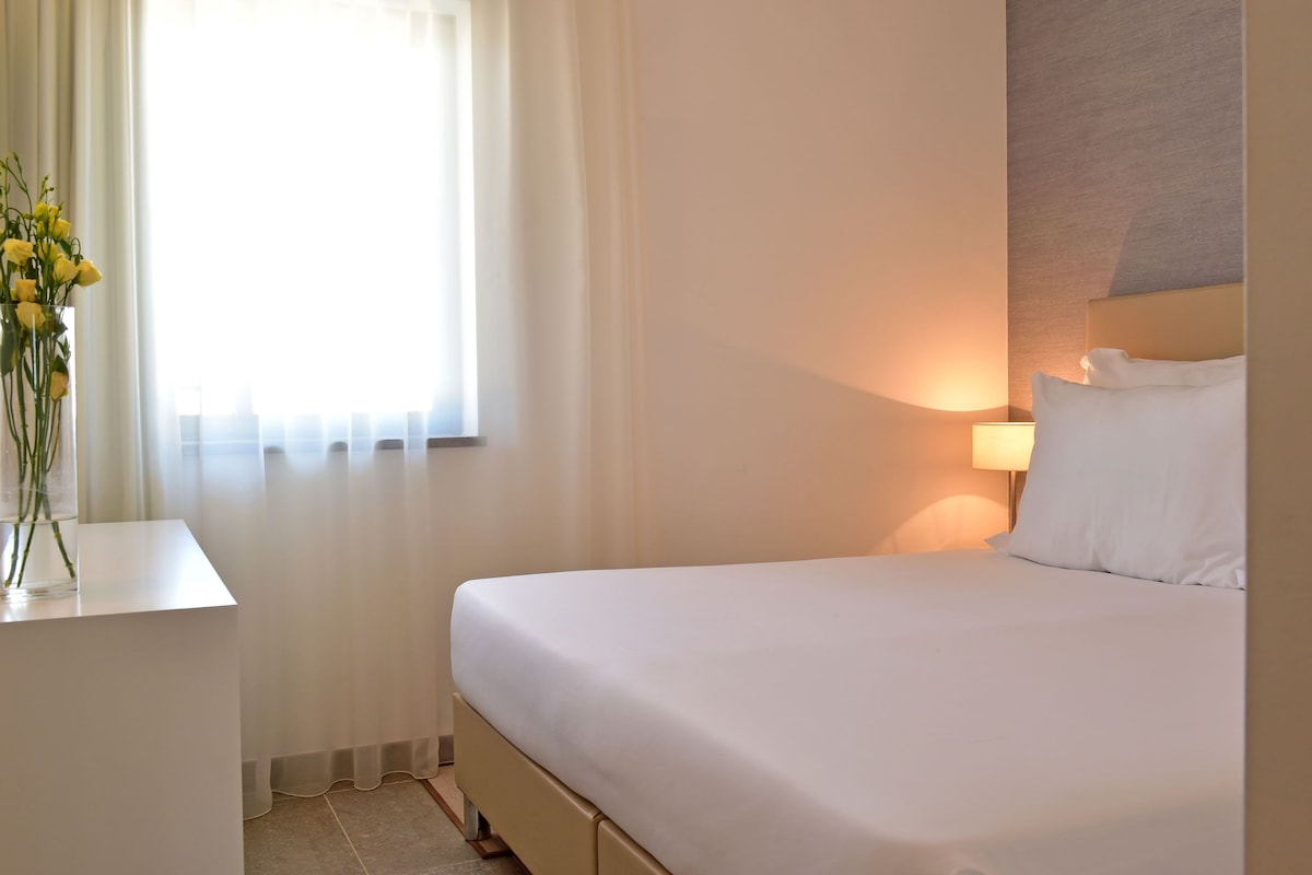 Una habitación de hotel con una cama, una mesita de noche con un jarrón de flores, una lámpara y una ventana con cortinas translúcidas.