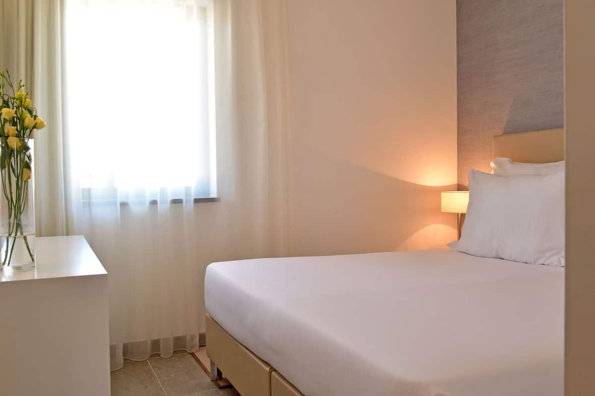 Une chambre d’hôtel avec un lit, une table de chevet ornée d’un vase de fleurs, une lampe et une fenêtre avec des rideaux légers.