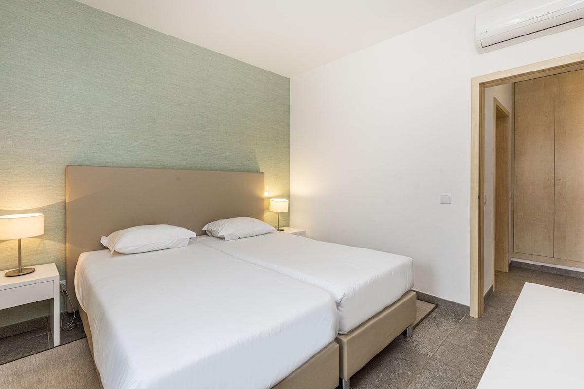 Une chambre d’hôtel avec deux lits simples, une table de chevet avec une lampe, une porte en bois et un climatiseur fixé au mur.