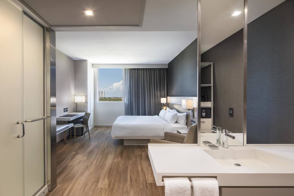 Une chambre d’hôtel moderne dotée d’un grand lit, d’un bureau, d’une salle de bain avec un grand miroir et offrant une vue sur la ville.