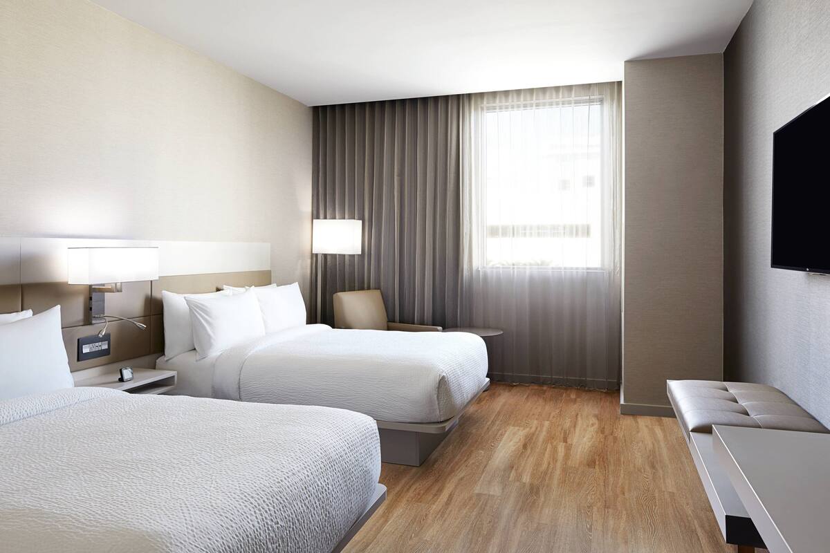 Une chambre d’hôtel avec deux lits, un téléviseur, une table de chevet avec une lampe, une chaise et une grande fenêtre avec des rideaux.