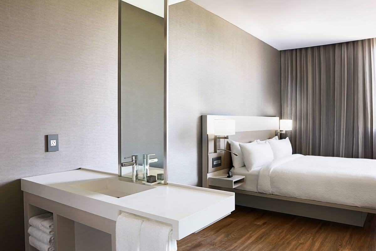 Une chambre d’hôtel moderne avec un grand lavabo blanc, un miroir, une table de chevet avec une lampe et un lit doté d’une tête de lit.
