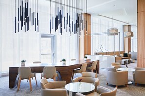A modern hotel lobby with a large wooden reception desk, comfortable seating, and a large window offering a city view.