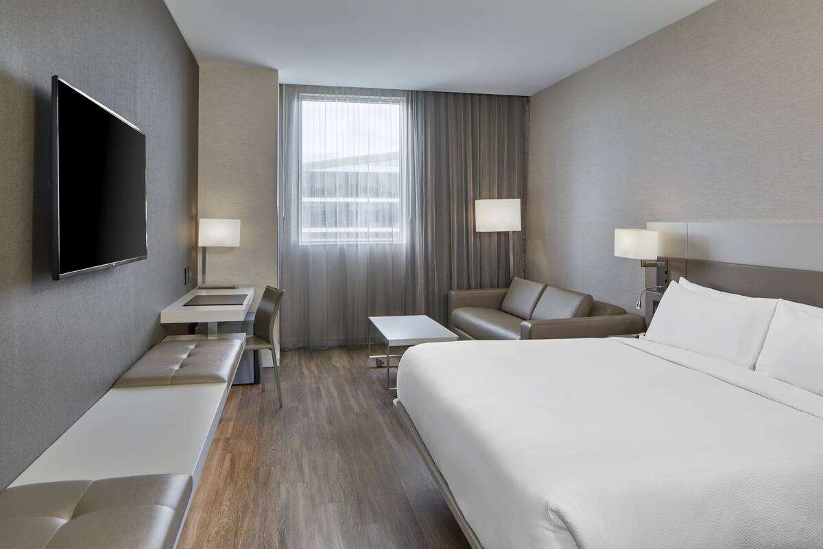 Une chambre d’hôtel moderne dotée d’un grand lit, d’un téléviseur à écran plat fixé au mur, d’un coin salon avec un canapé et des fauteuils, et d’une petite table.