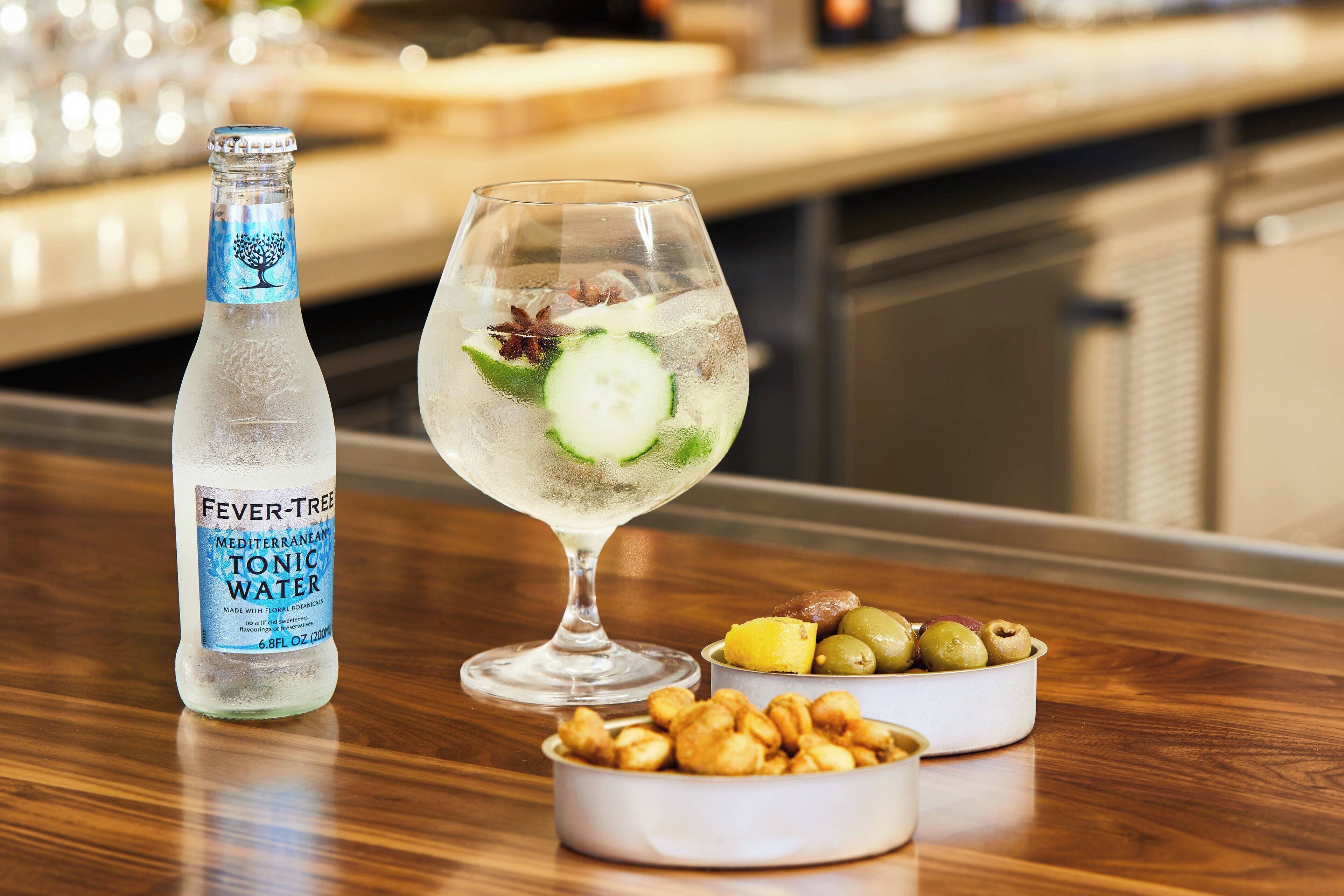 Una botella de Fever-Tree Tónica Mediterránea, un vaso de ginebra con pepino y menta, y un cuenco con frutos secos y aceitunas sobre una mesa de madera.