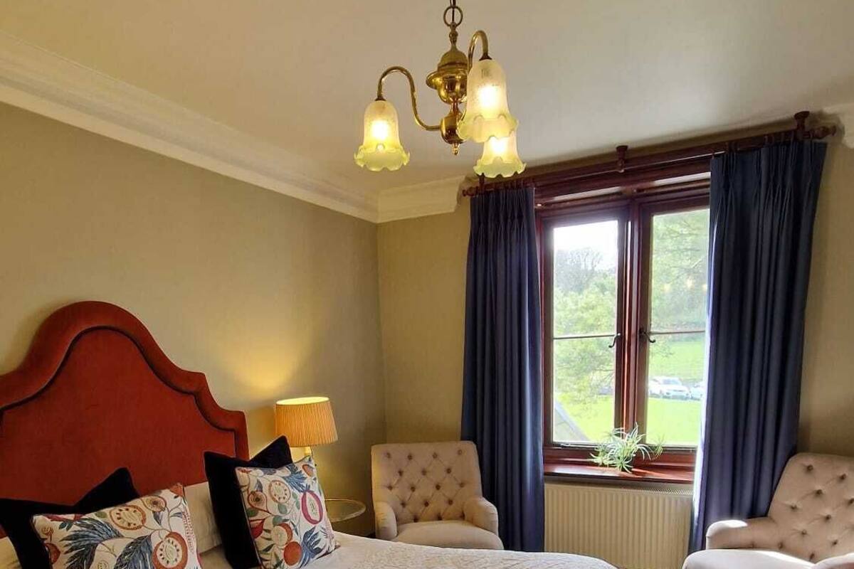 A spacious bedroom with a large bed, two armchairs, a wooden headboard, a chandelier, and a window with curtains.
