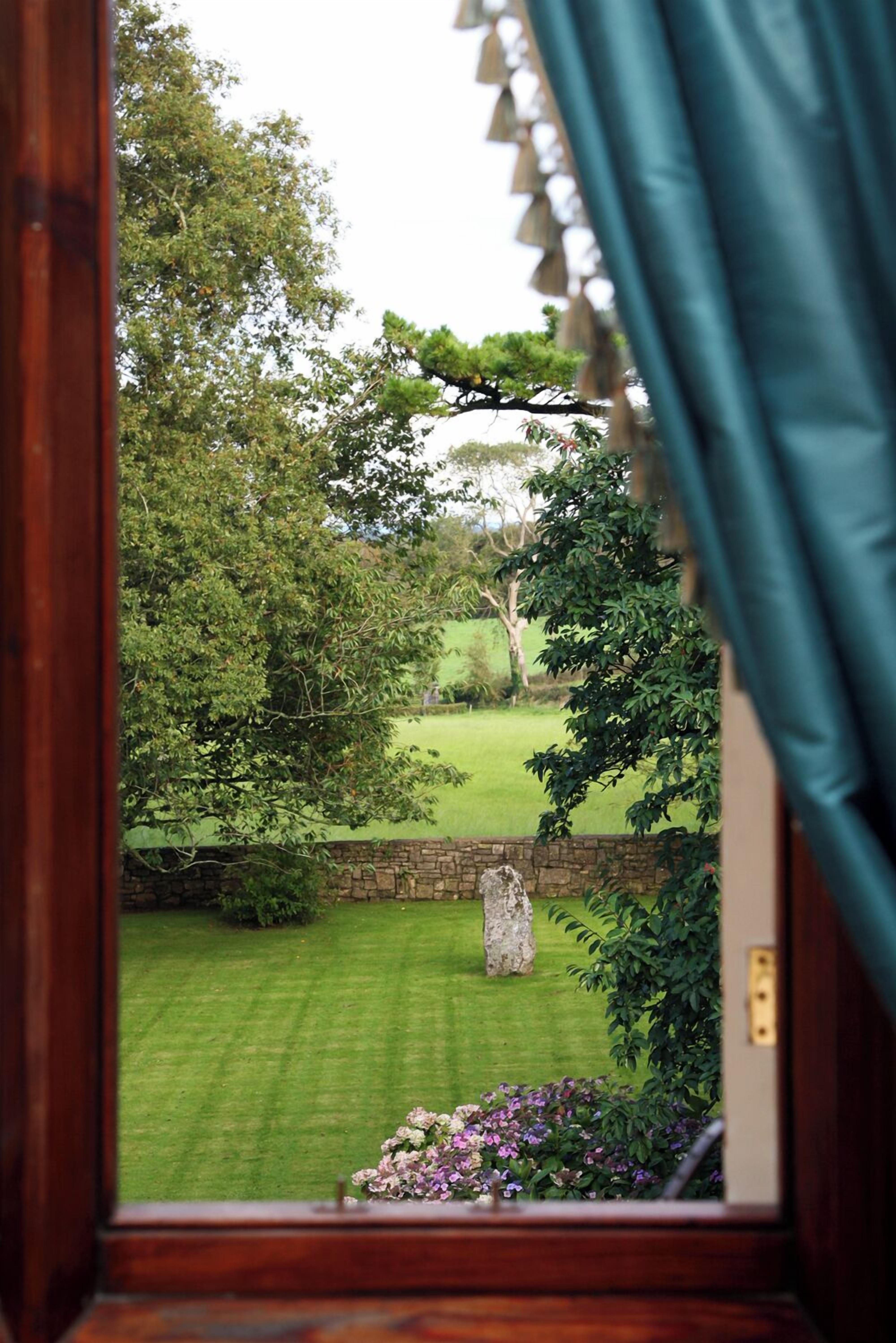 A view through a window with a stone wall and a grassy garden with trees.