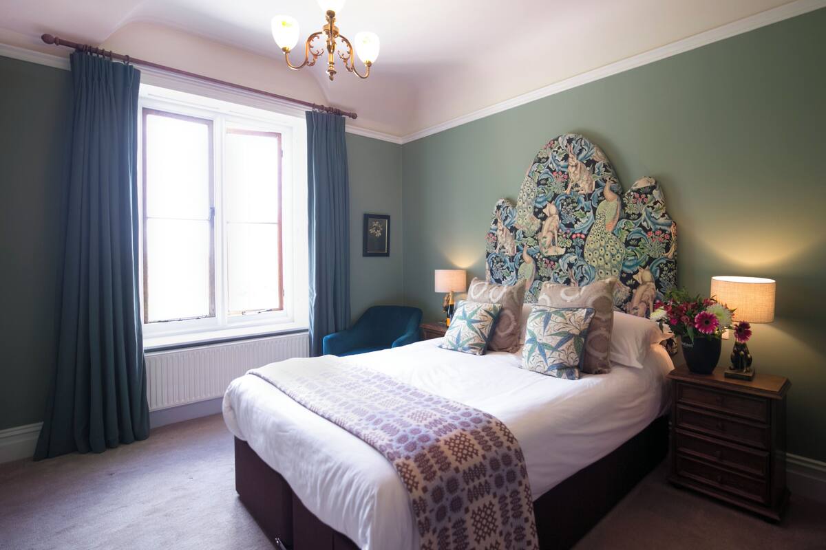 A bedroom with a large bed, a nightstand with a lamp, a chair, and a window with curtains.