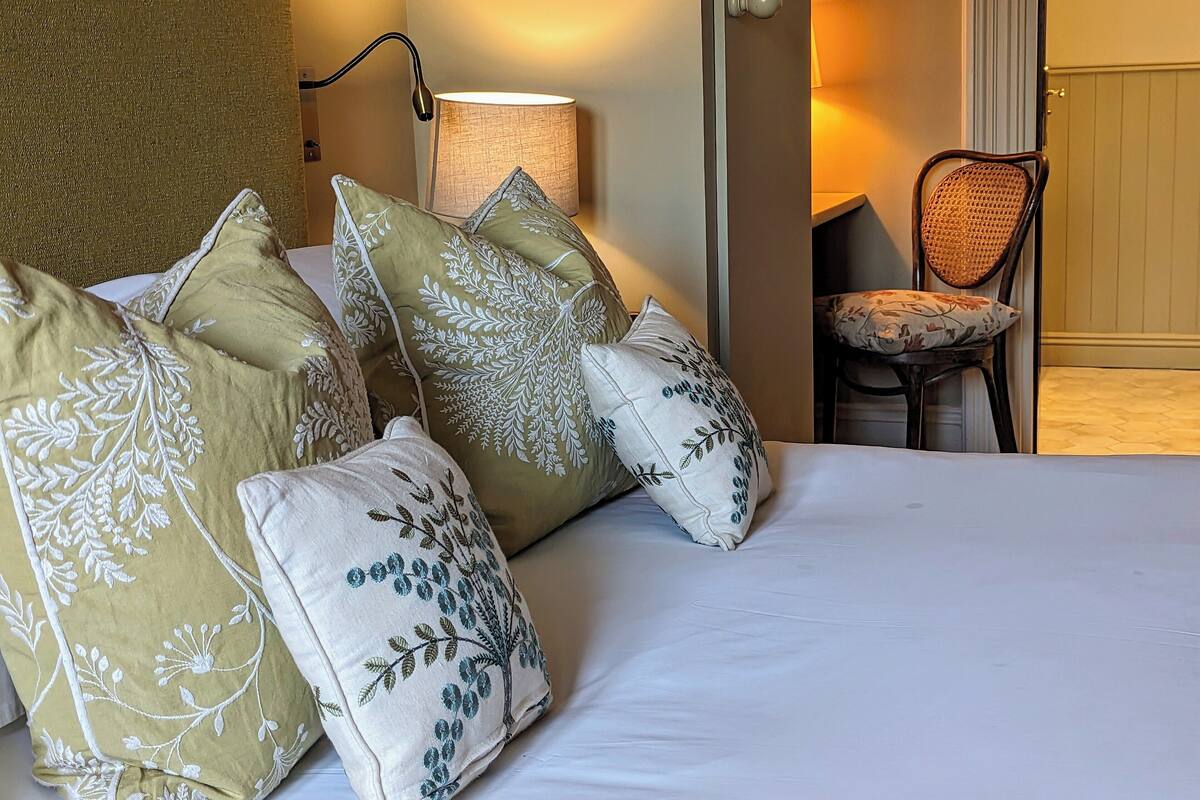 A neatly made bed with several decorative pillows, a bedside lamp, and a chair in the background.