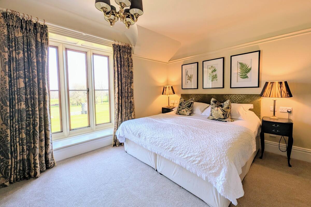 A well-lit bedroom with a large bed, two bedside tables, a chandelier, and framed botanical prints on the wall.