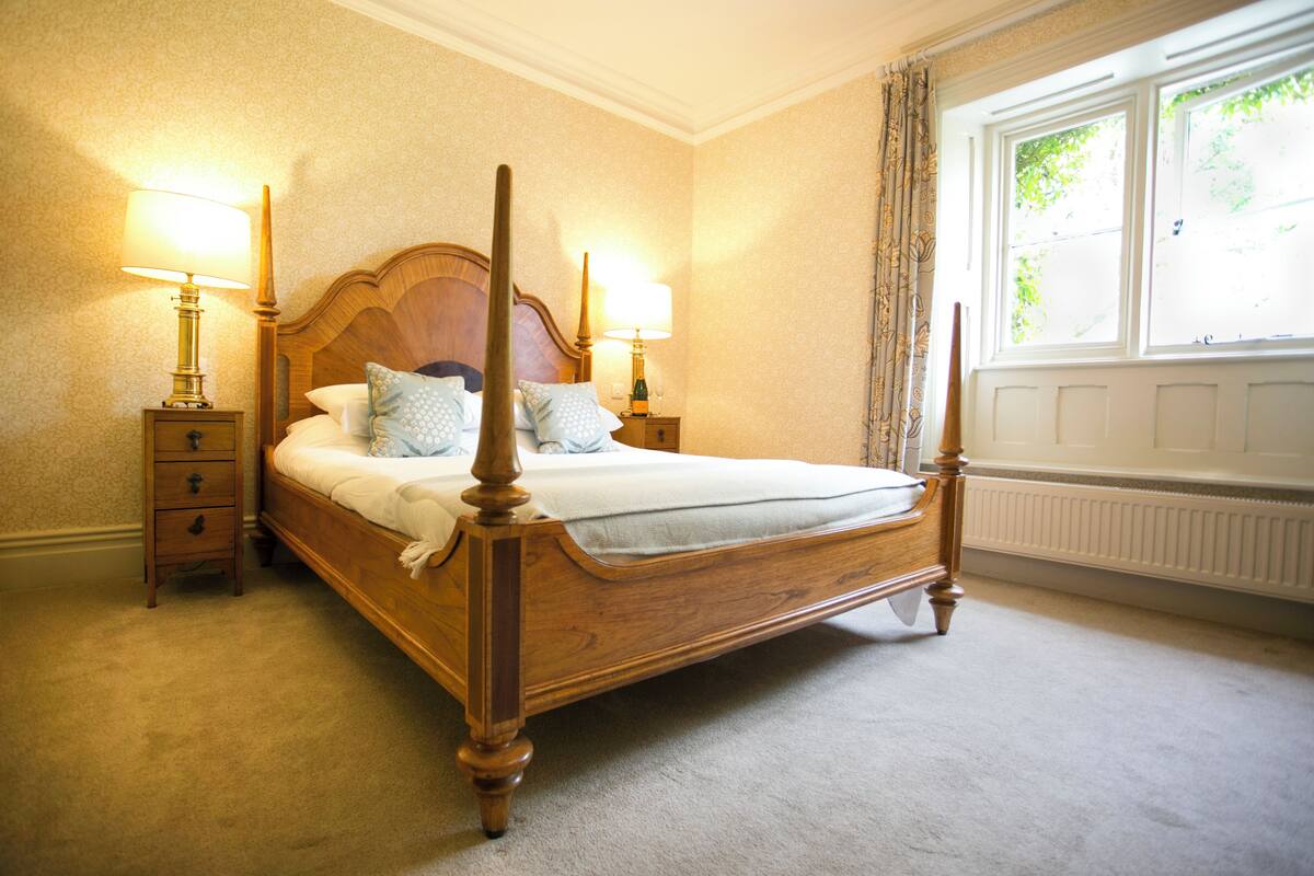 A four-poster bed with two bedside tables, each with a lamp, in a room with a radiator and a window with curtains.