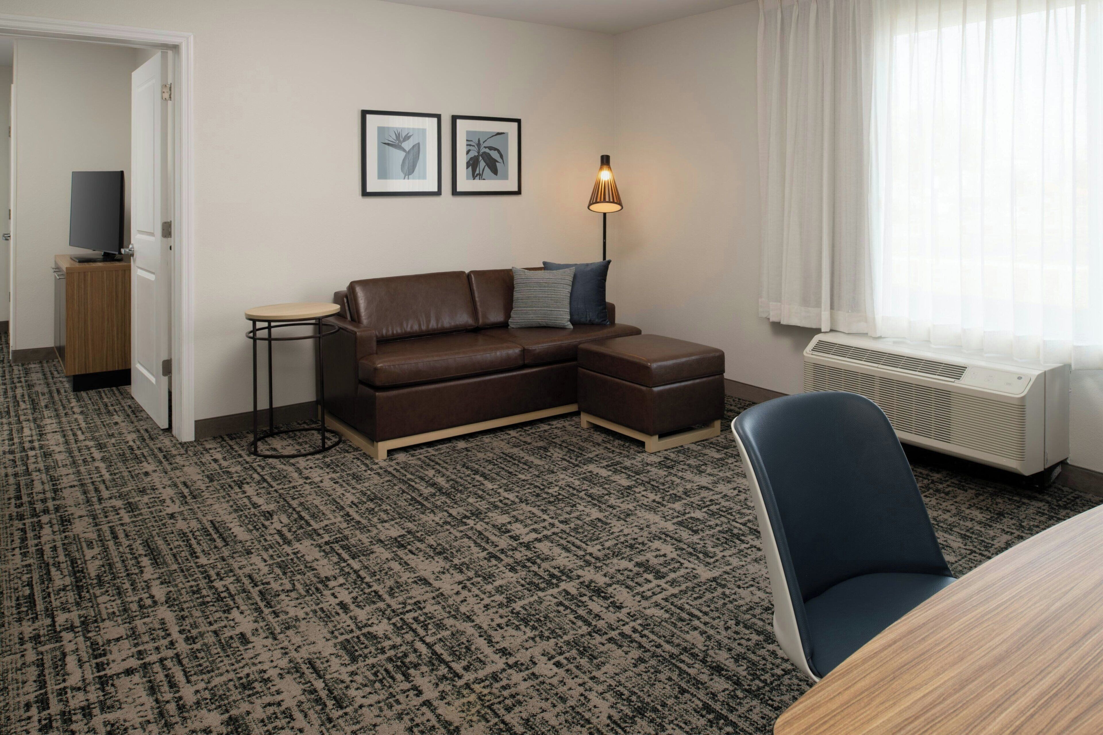 Habitación de hotel moderna con un sofá seccional marrón, una mesita auxiliar pequeña, una silla azul, una lámpara de pie y un ventanal con cortinas traslúcidas.
