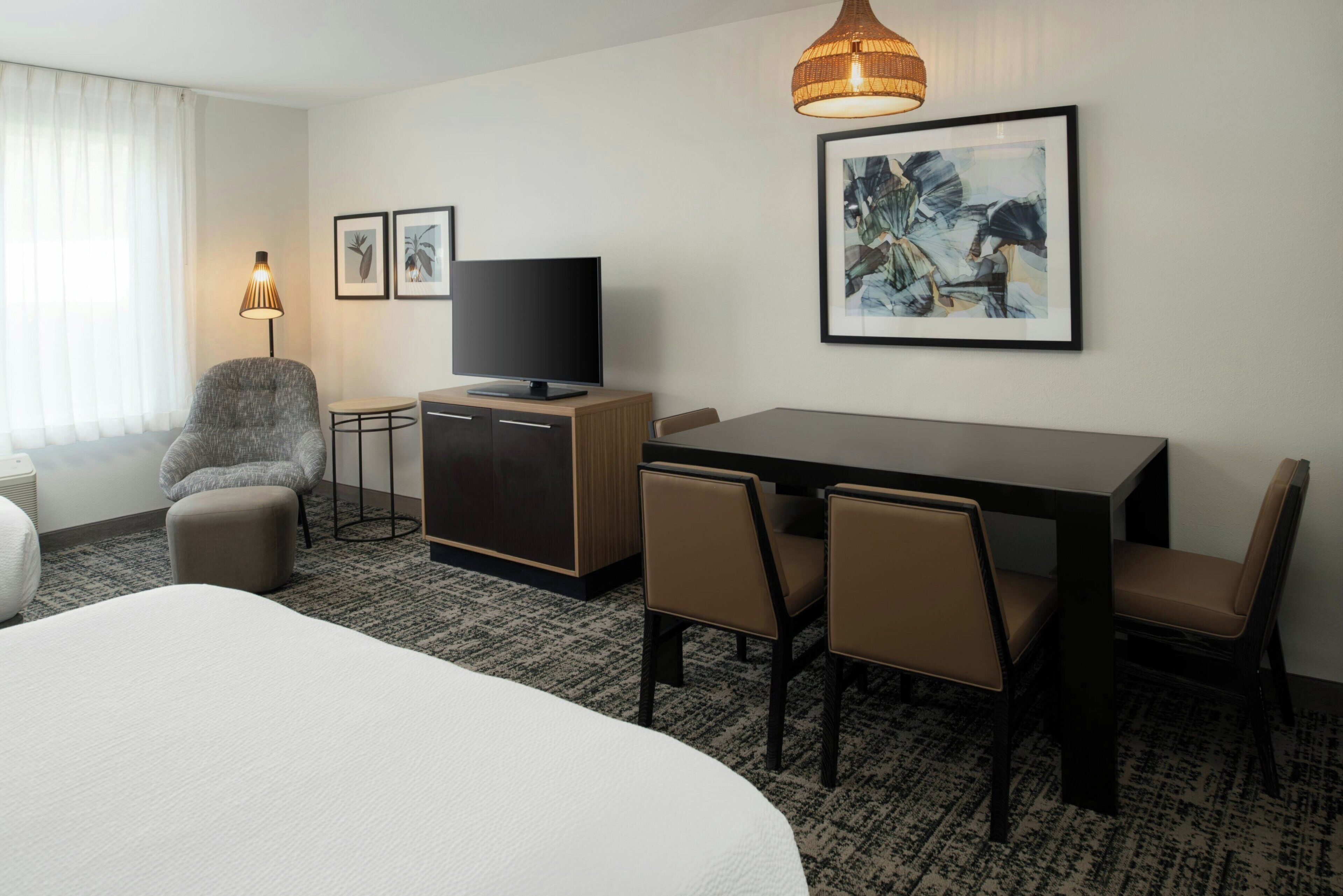 Quarto de hotel com cama, escrivaninha com televisão, cadeira e mesa de jantar com cadeiras.