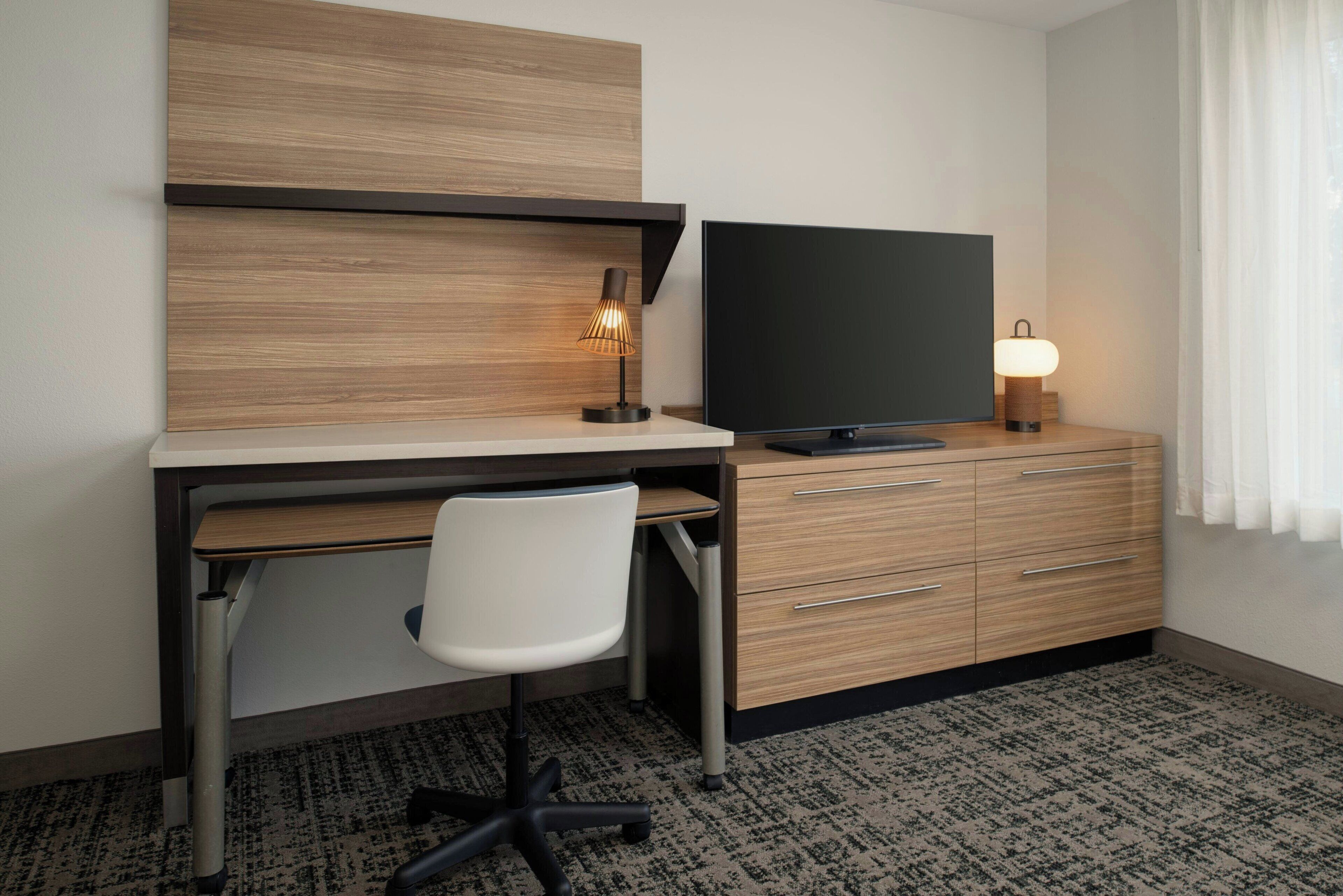 Une chambre d’hôtel avec un bureau en bois, une chaise moderne, un téléviseur à écran plat et une table de chevet avec une lampe.