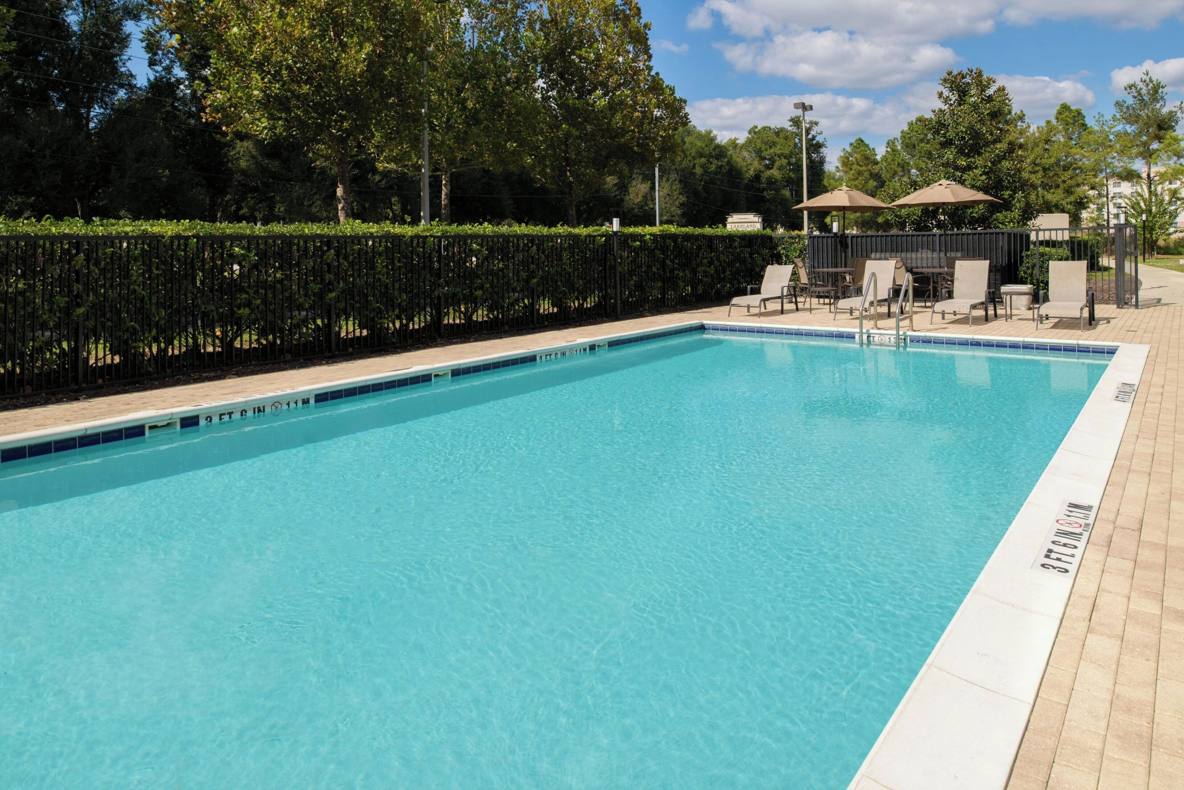 A rectangular swimming pool with clear blue water, surrounded by a tiled deck. There are lounge chairs and umbrellas in the background.