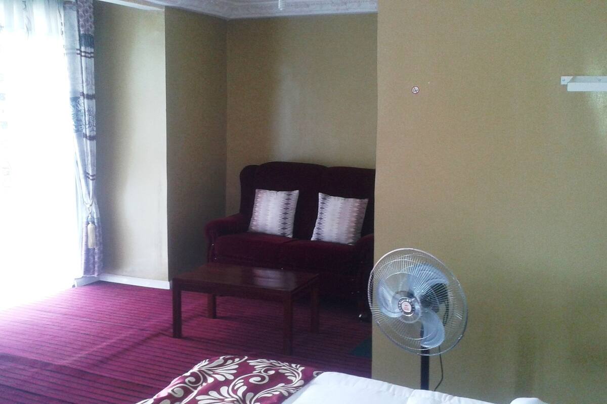 A hotel room with a red armchair, a small table, and a standing fan.